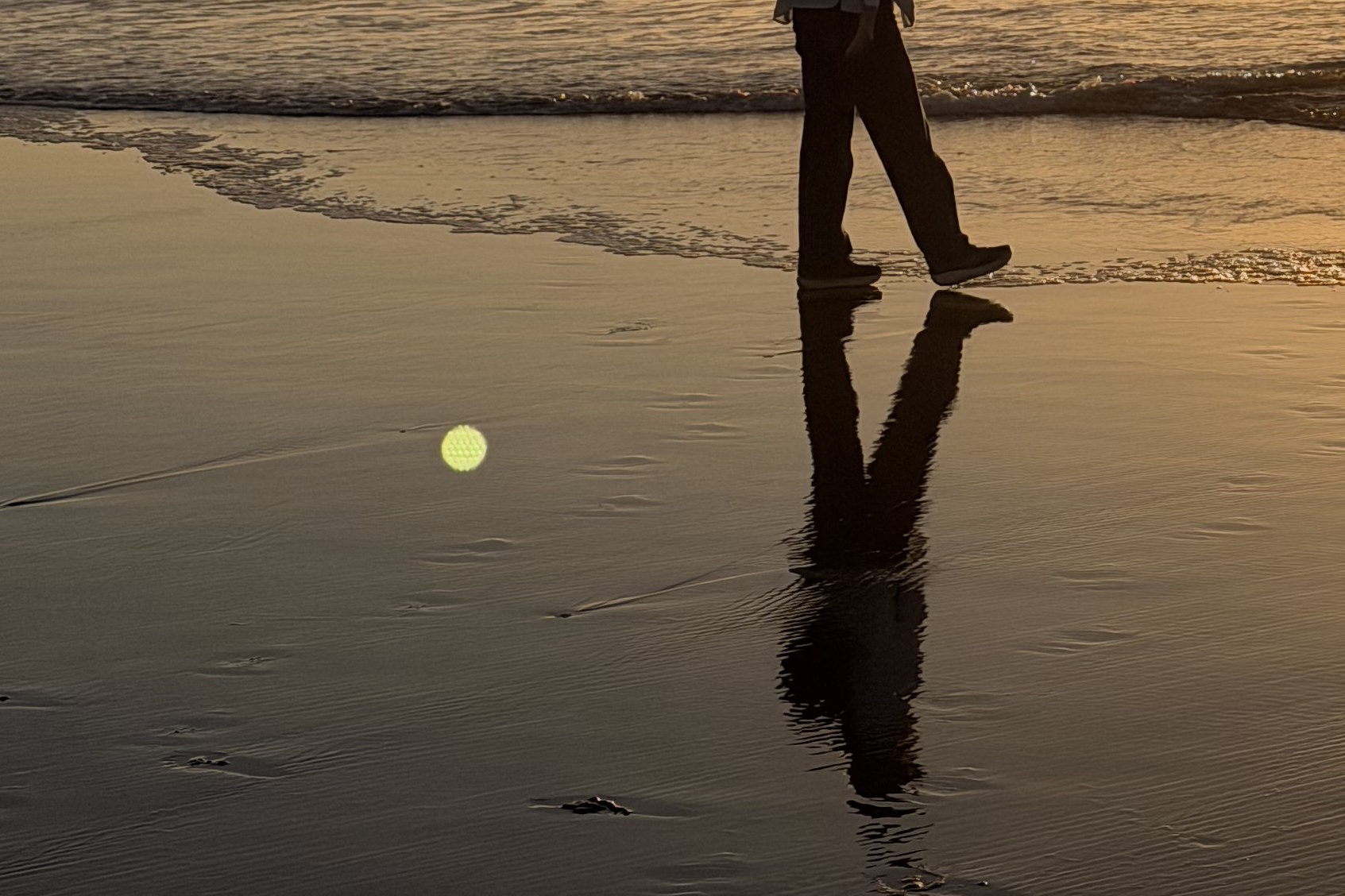 Wet sand at sunset with a walker's reflection and a small green lens ghost on the left